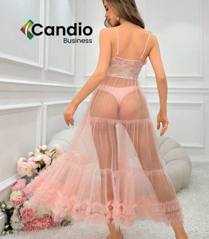 Une femme se tient dos à la caméra, vêtue de la Robe de nuit en maille, un ensemble de lingerie rose transparent mettant en valeur une longue jupe fluide superposée. Elle est installée dans une pièce de couleur claire avec des sièges moelleux et des compositions florales. Le texte « Candio Business » est visible dans le coin supérieur gauche.